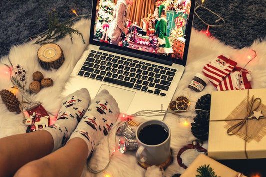 Person in Christmas socks watching a holiday movie on a laptop, surrounded by gifts, decorations, and a mug of coffee.